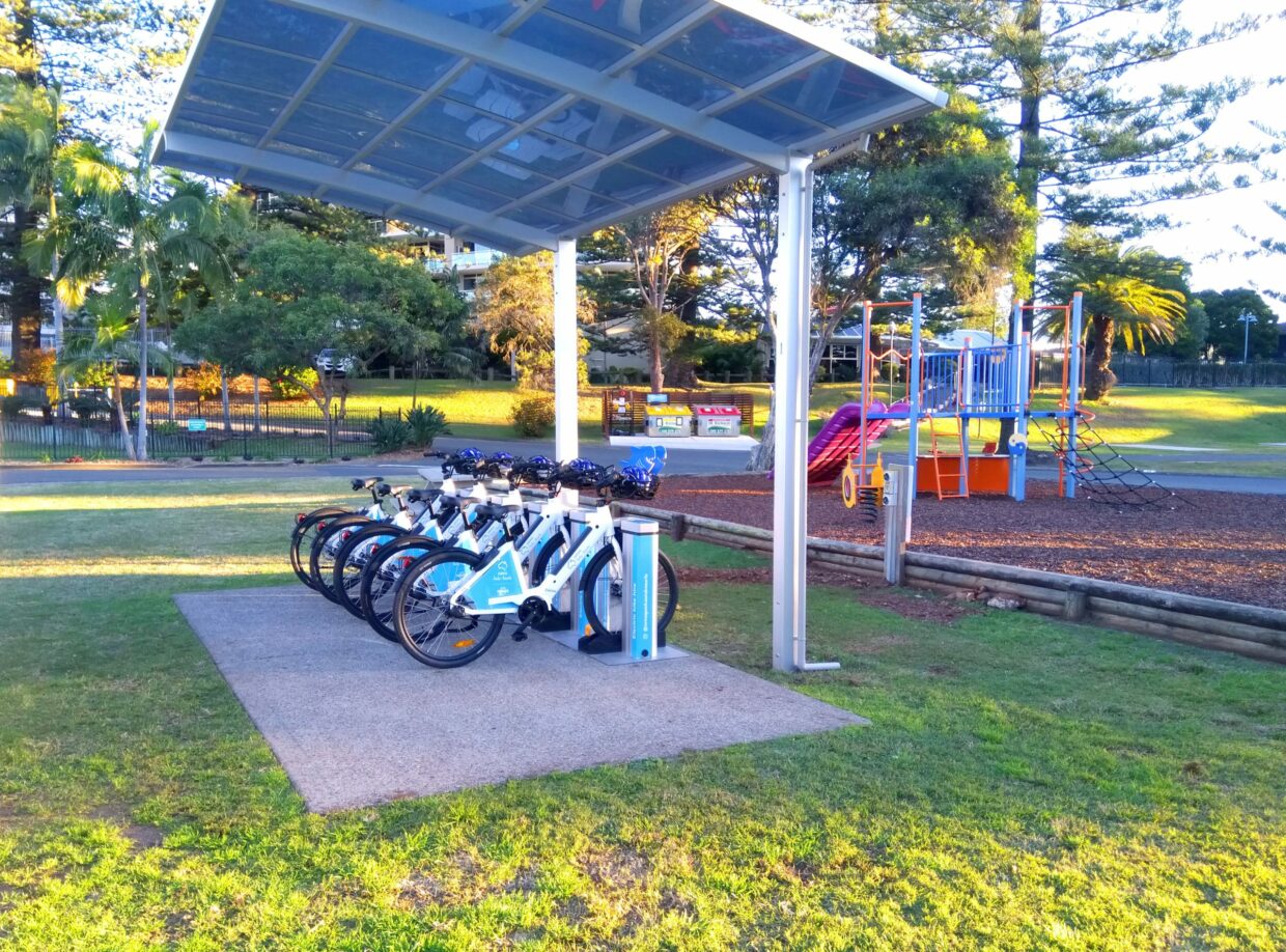 A covered BYKKO e-bike charging station with white e-bikes docked in an Australian holiday park, beside a children's playground.