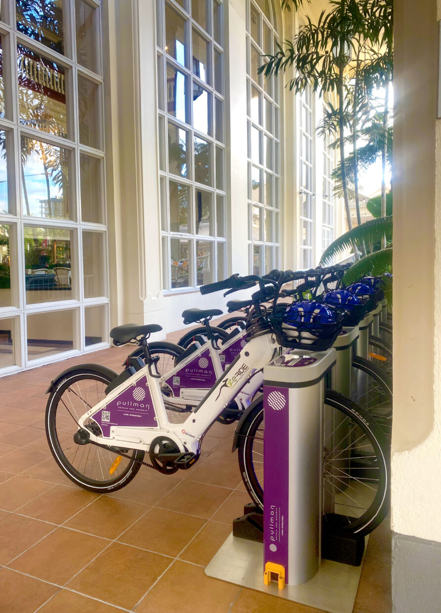 A row of white BYKKO e-bikes docked and locked at a charging station in a Pullman hotel lobby, helmets in the baskets.