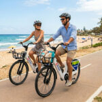 A smiling couple ride white BYKKO e-bikes along a coastal shared path beside a beach on a sunny day.