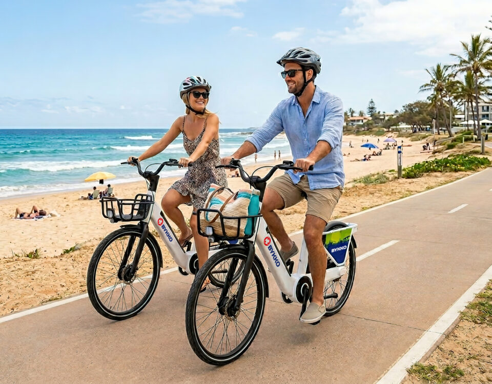 A smiling couple ride white BYKKO e-bikes along a coastal shared path beside a beach on a sunny day.
